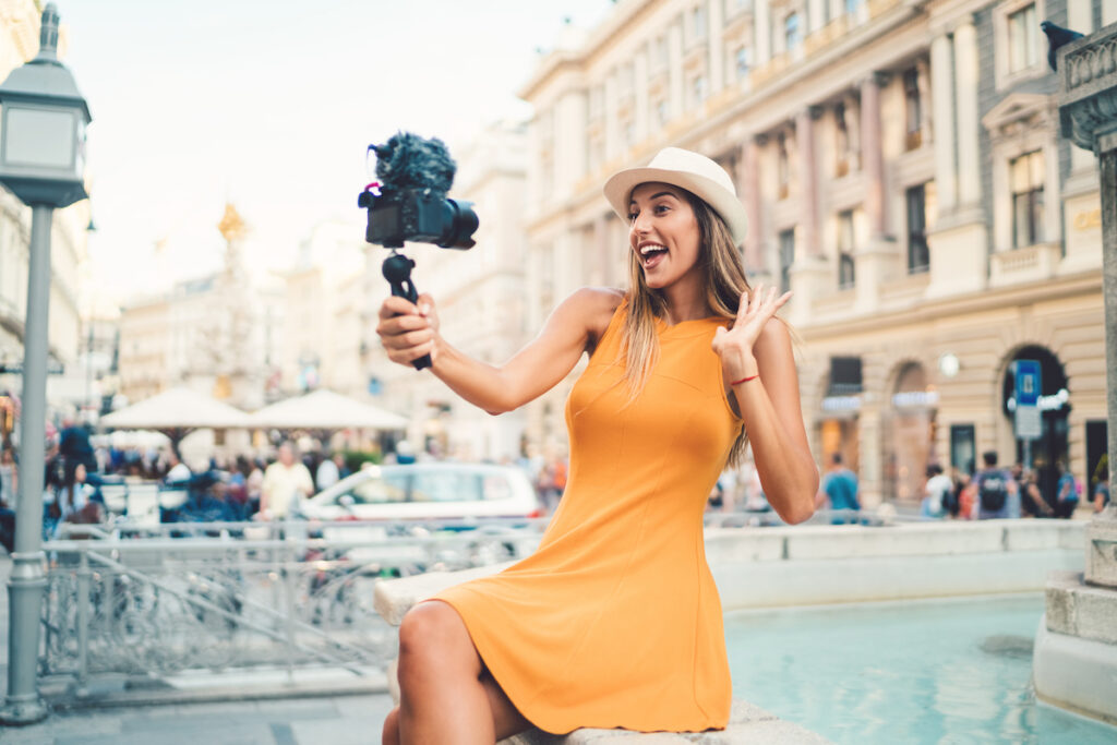 Eine Influencerin posiert auf dem Rand eines Brunnens, hält eine Kamera in der rechten Hand und winkt mit der linken Hand in die Kamera.